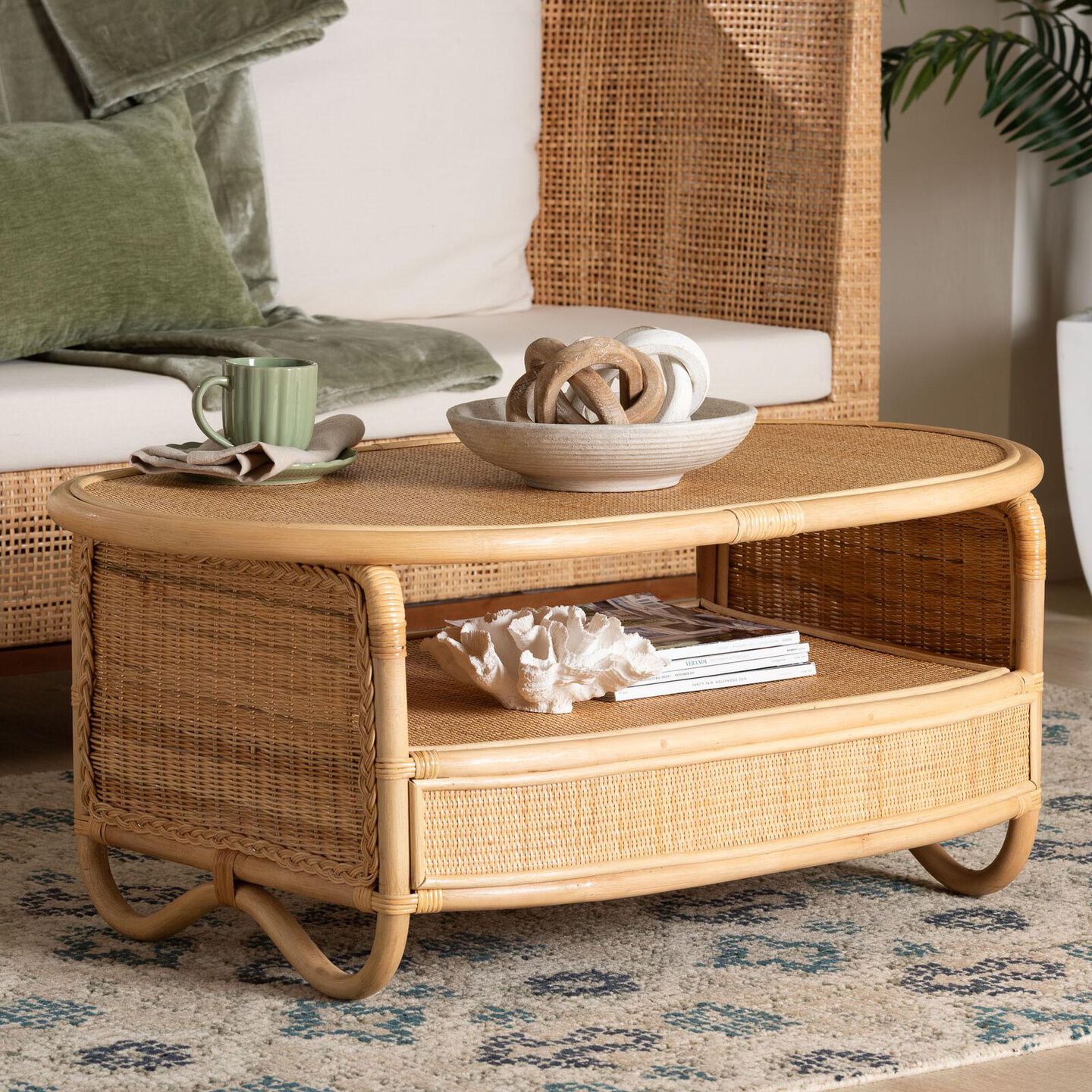Eclectic rattan coffee table with neutral toned accents - a bowl with w whimsical objects and a coffee cup and saucer on top of the table. A natural seashell bowl and stacked books in the middle open area.