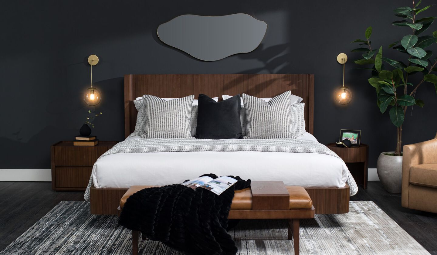 Modern bedroom with walnut wood headboard, black accent pillows, brass wall sconces, and a stylish armchair.