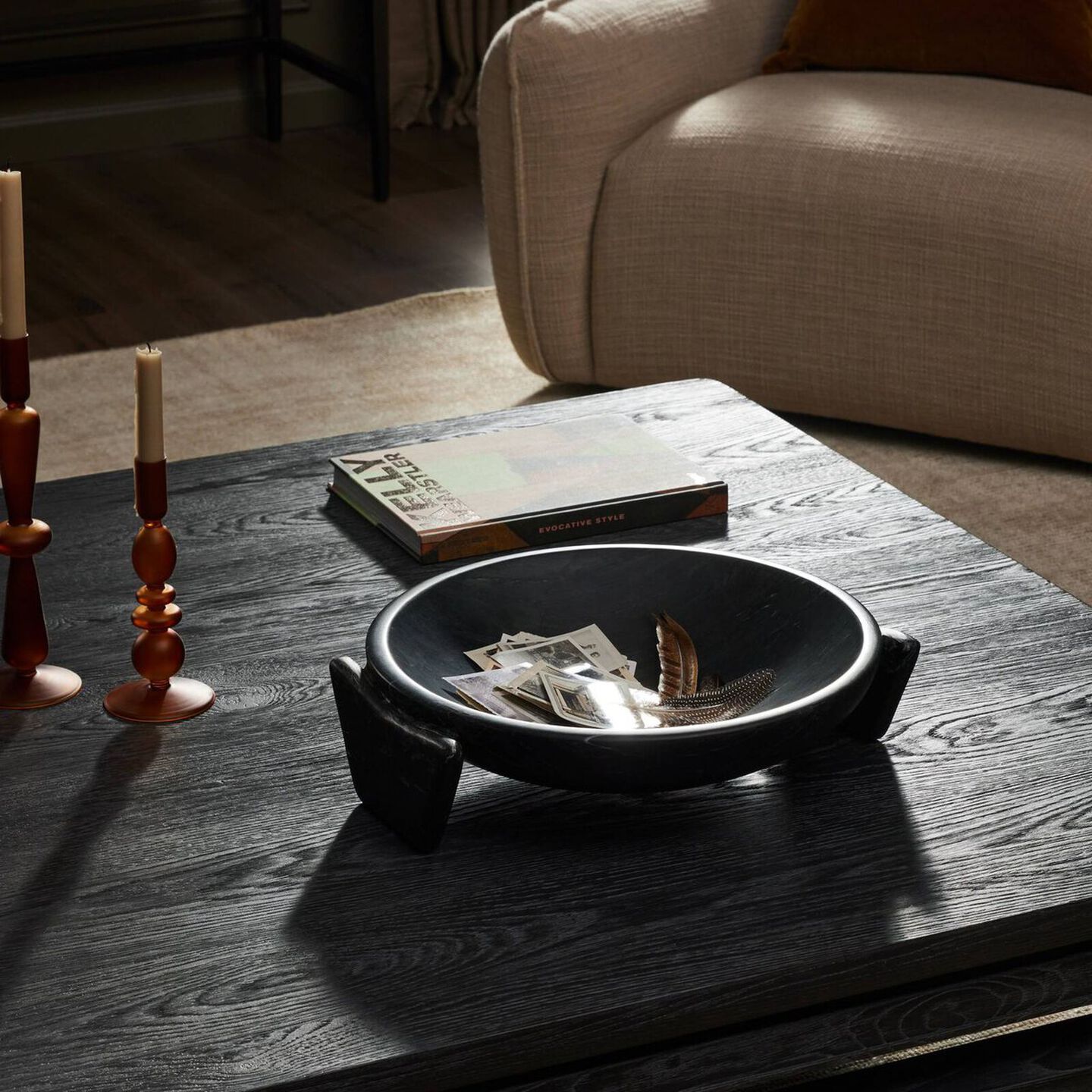 A wide shallow bowl that appears to float effortlessly between two soft-edged, cradle-style legs styled on a coffee table with two decorative candlesticks and a decorative book.